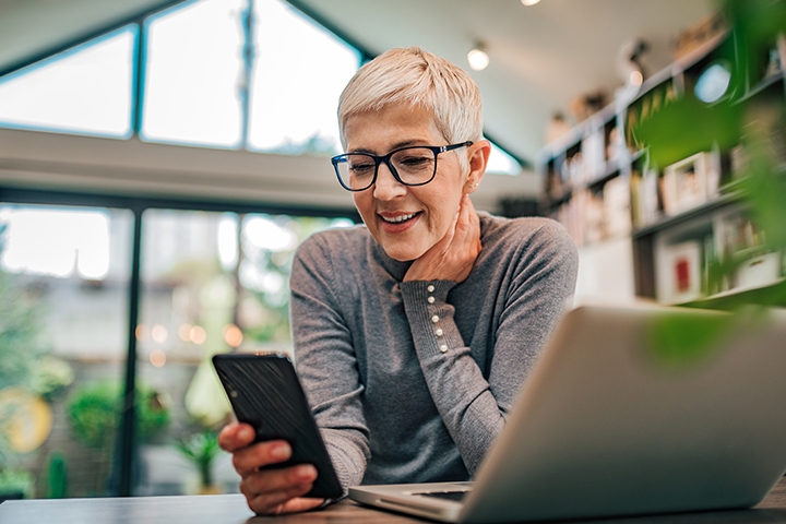 Frau mit Brille vor dem Laptop mit dem Handy in der Hand