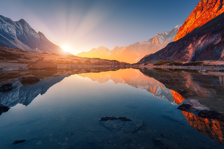 Beautiful landscape with high mountains with illuminated peaks, stones in mountain lake, reflection, blue sky and yellow sunlight in sunrise.