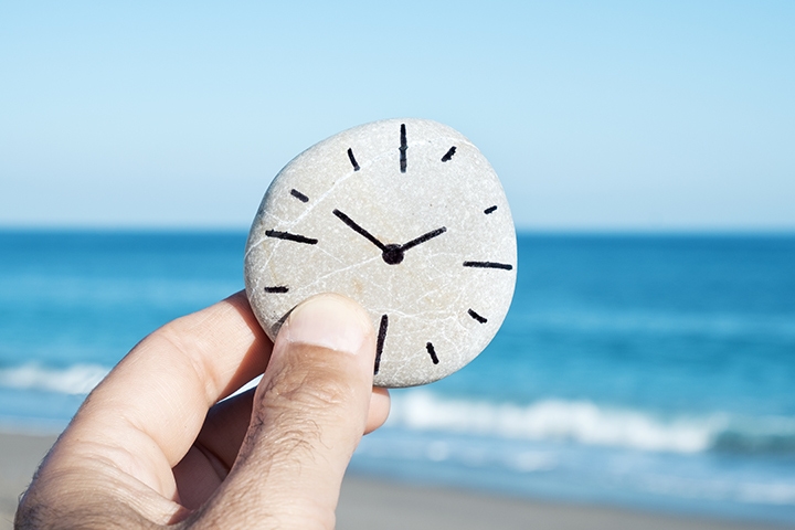 Person holding rock with hand-drawn clock on it in front of the ocean on a sandy beach