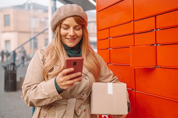 Woman standing in front of parcel locker holding phone and package