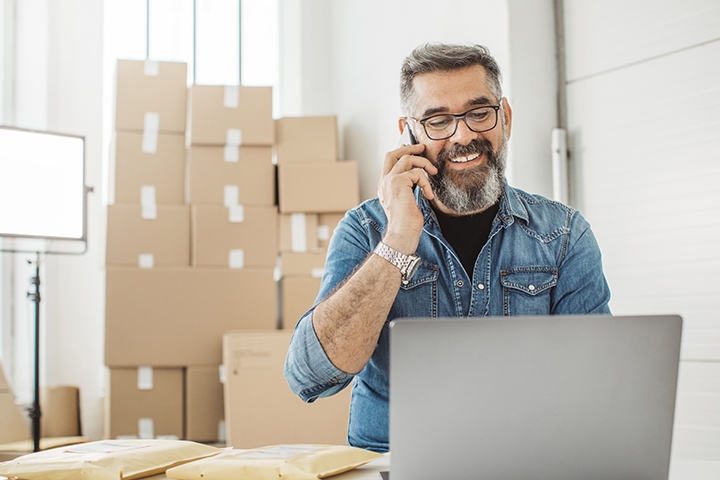 Man with beard and glasses on phone in front of pile of box packages