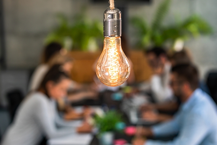 Bright lightbulb hanging in the forefront while blurred in the background is a group of young people working together on a project