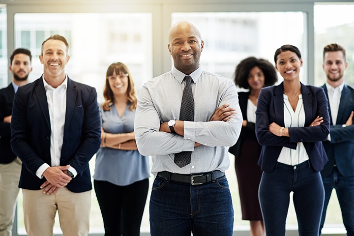 Professional team standing together in an office, with a man in the foreground holding documents while colleagues stand behind him.