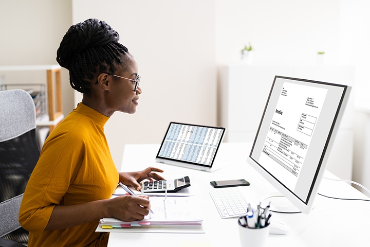 A woman sits at a desk working on a computer monitor that displays charts and data. A tablet is open beside her with additional documents. She appears focused as she reviews the information in a bright, modern office setting.
