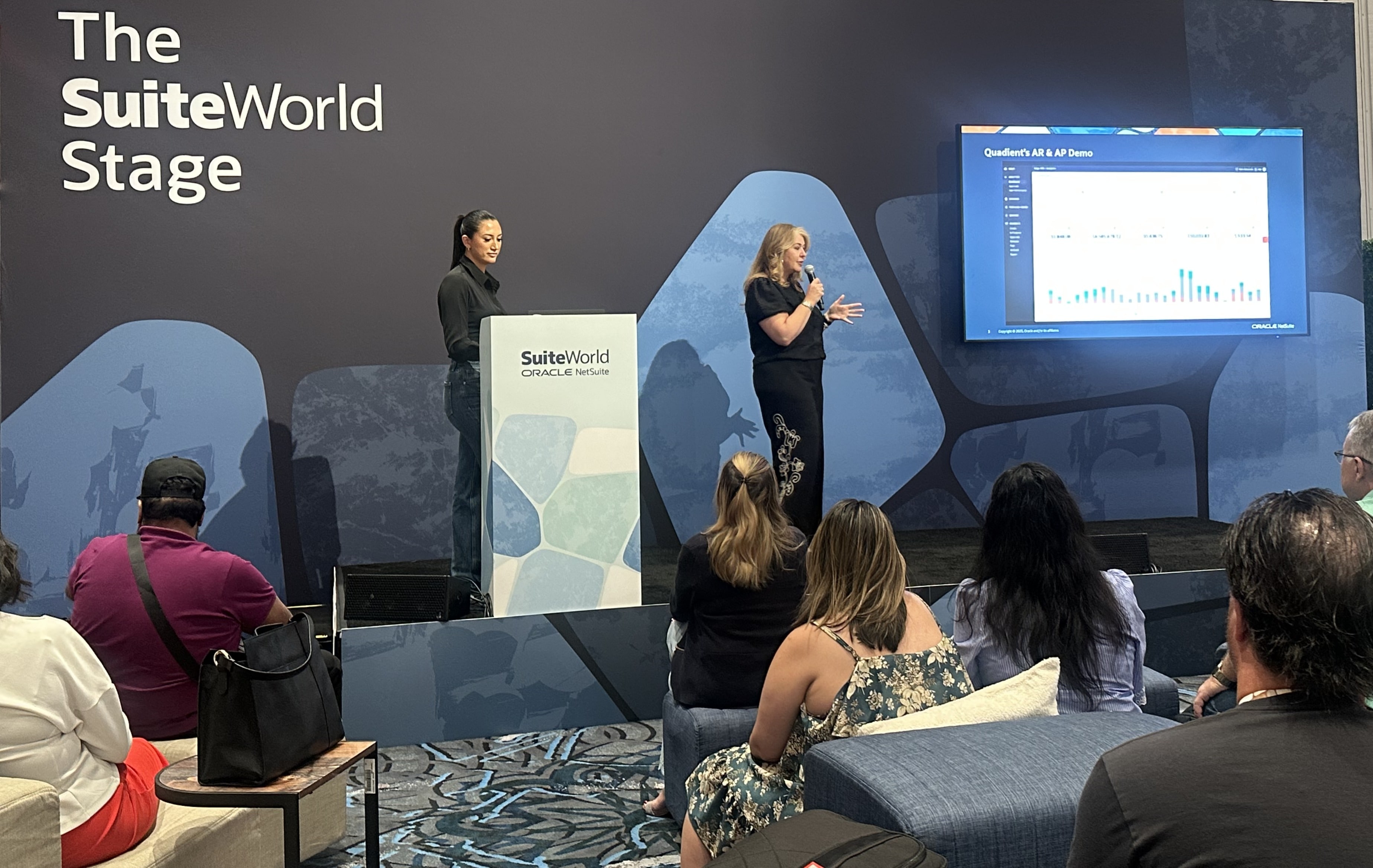Speaker presenting Quadient AP and AR automation demo on stage at SuiteWorld2025, with a large screen showing financial workflow charts and an audience watching.