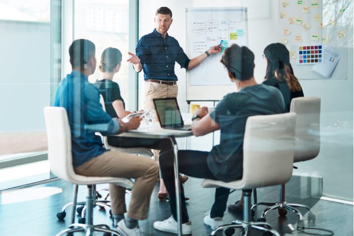 Team in a modern office collaborating during a presentation, with a person standing near a whiteboard covered in notes and diagrams.