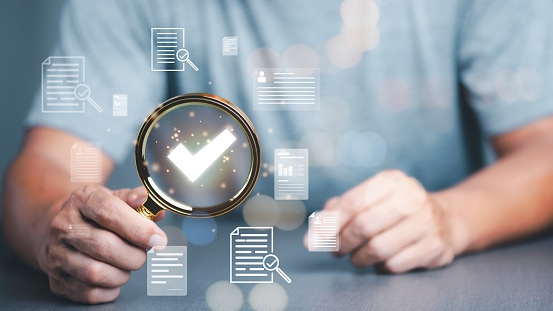 Hands holding a magnifying glass highlighting a checkmark icon, surrounded by floating digital document symbols representing document review, accessibility verification, and compliance checks.