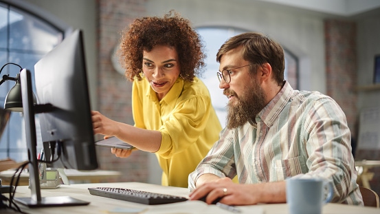 A woman points at a laptop screen while discussing something with a man who is seated at the desk.