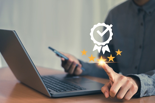 Person using a tablet and laptop with a digital compliance checkmark and stars overlay,.