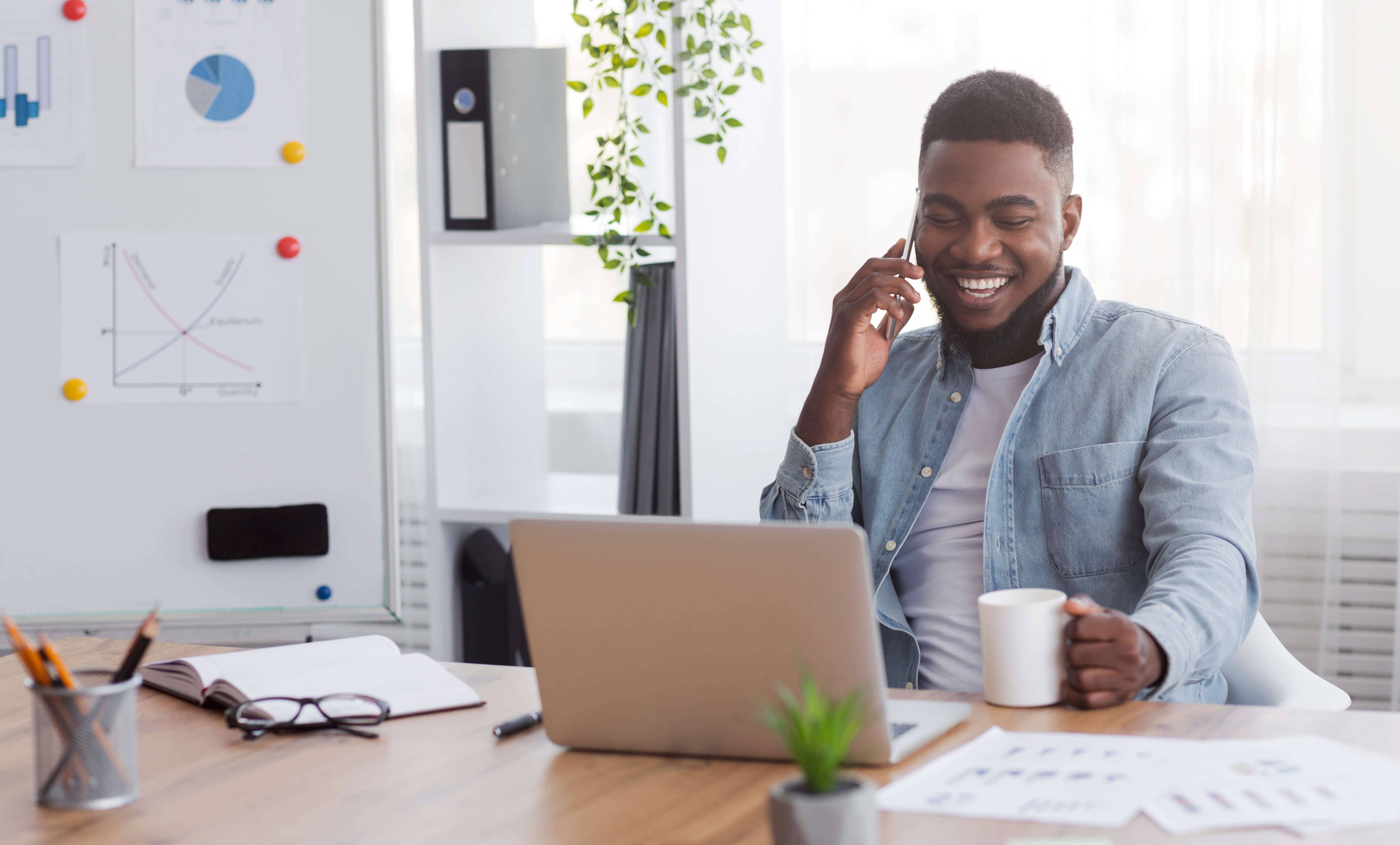 male talking on the phone with a cup of coffee and laptop on the desk