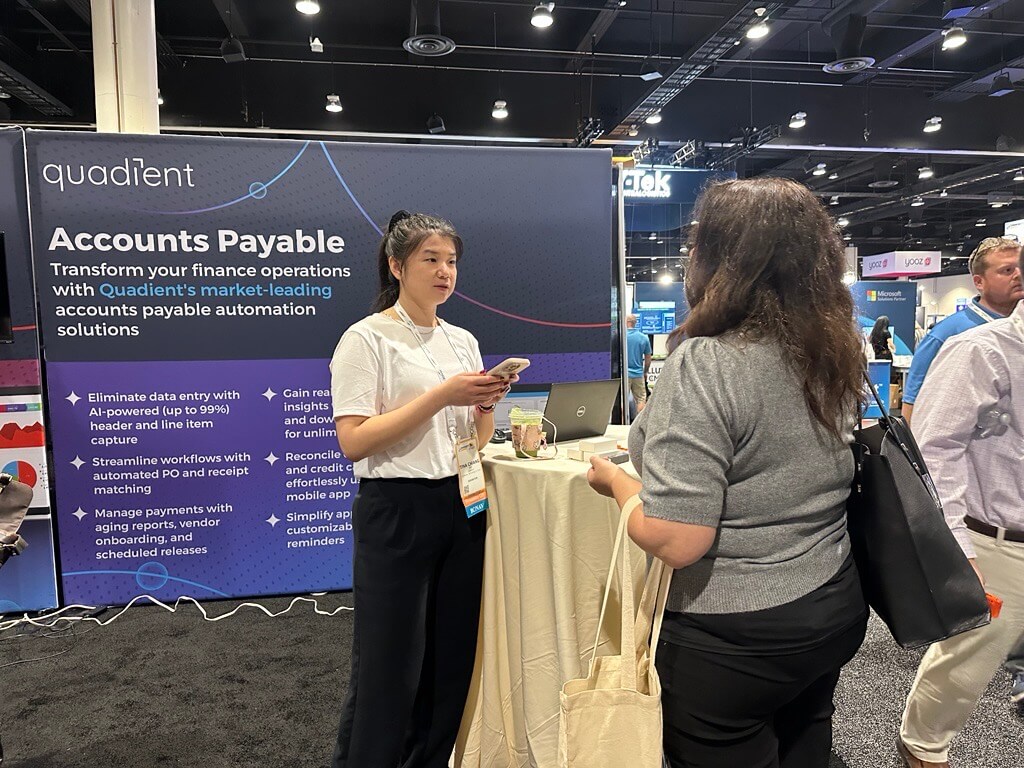 Two people are standing and conversing at a trade show booth for Quadient. The booth features a large banner that reads “Accounts Payable – Transform your finance operations with Quadient’s market-leading accounts payable automation solutions.” A high-top table with a laptop and coffee cup is in front of the banner. The background shows other booths and attendees in a convention hall.