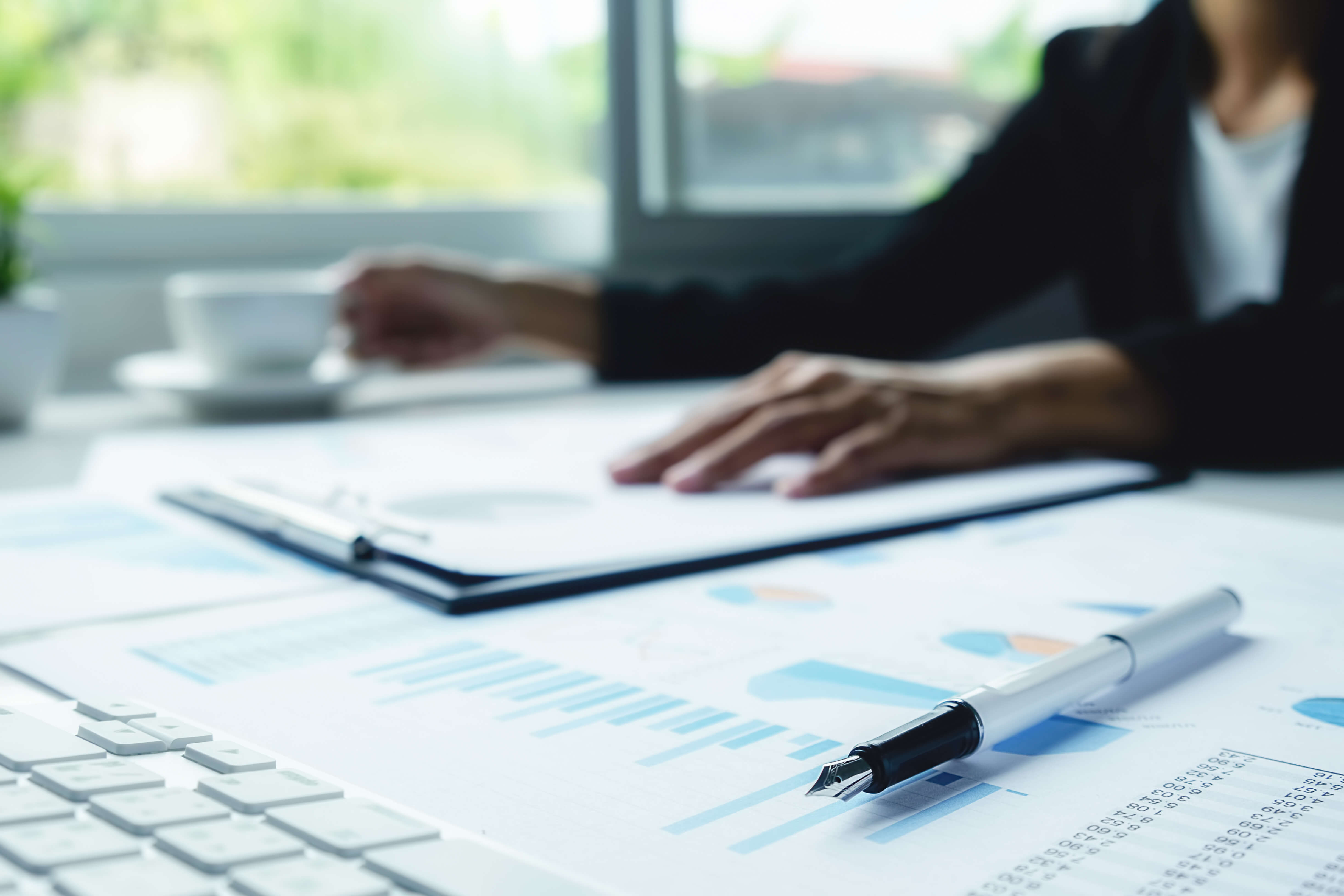 Close-up of financial documents with charts and graphs on a desk, a pen placed on top, and a person reviewing paperwork near a keyboard.