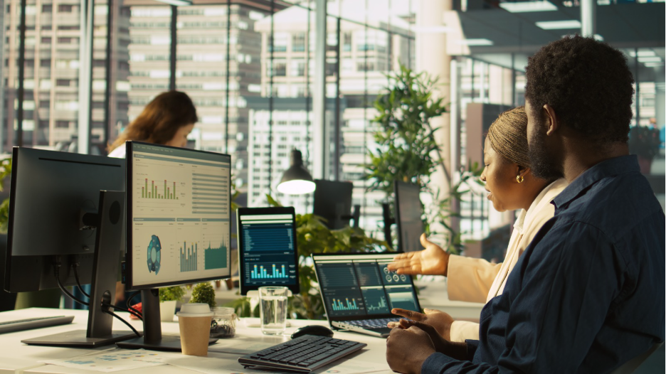 Person sitting at a desk reviewing printed documents in front of a computer screen and printer in an office setting