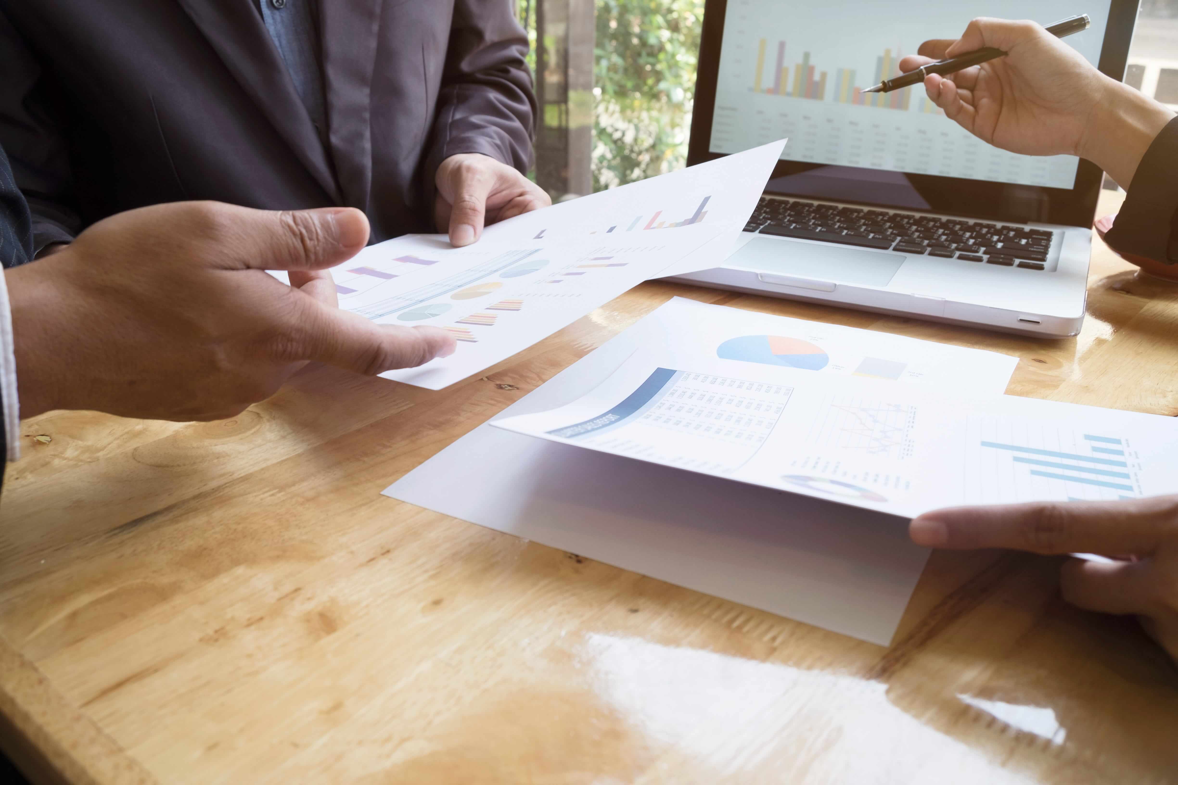 Close-up of hands holding printed financial reports with charts and metrics, with a laptop displaying similar analytics, representing analysis work involved in optimizing AP and AR.
