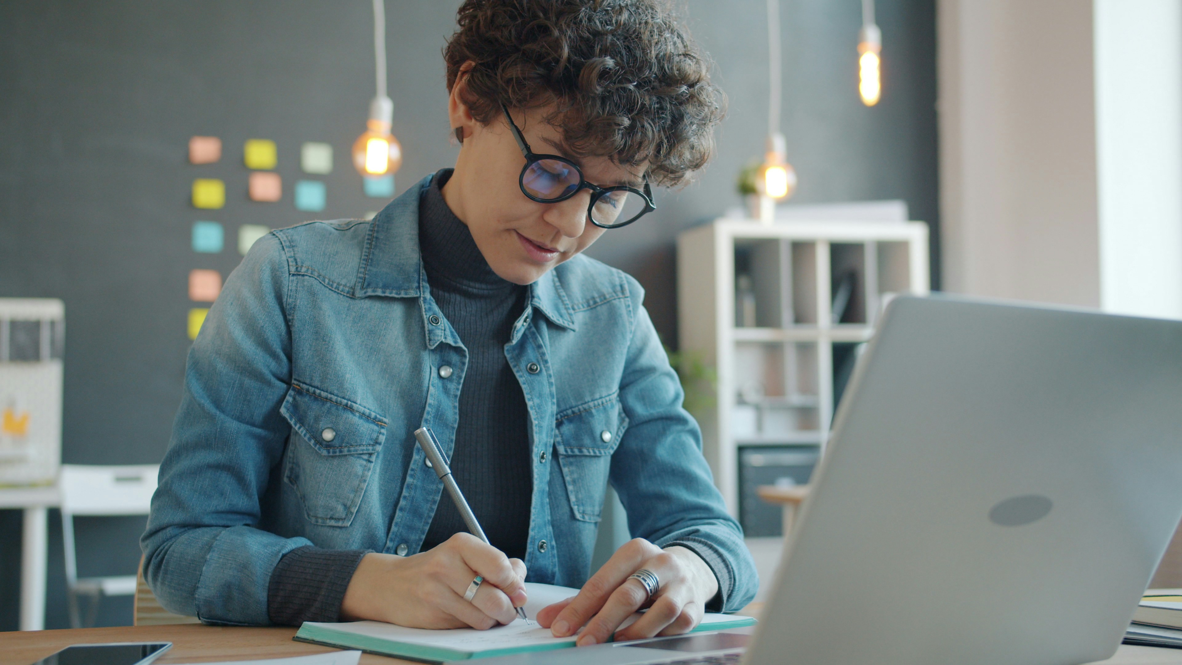 Person working at a desk with a laptop, writing notes in a notebook in a modern office setting